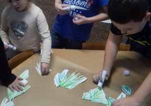 troje dzieci przy stole przykleja wycięte elementy choinek na szarym papierze